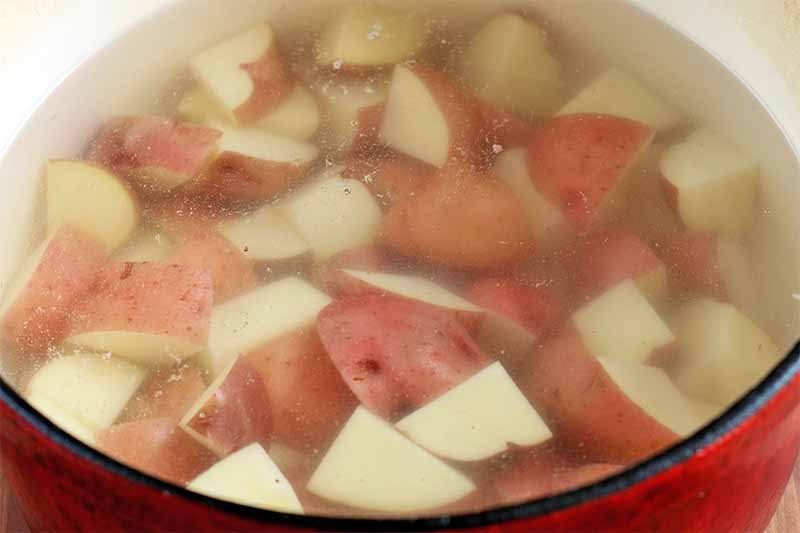 A red enameled pot of water with red-skinned potatoes cut into chunks, ready to boil.