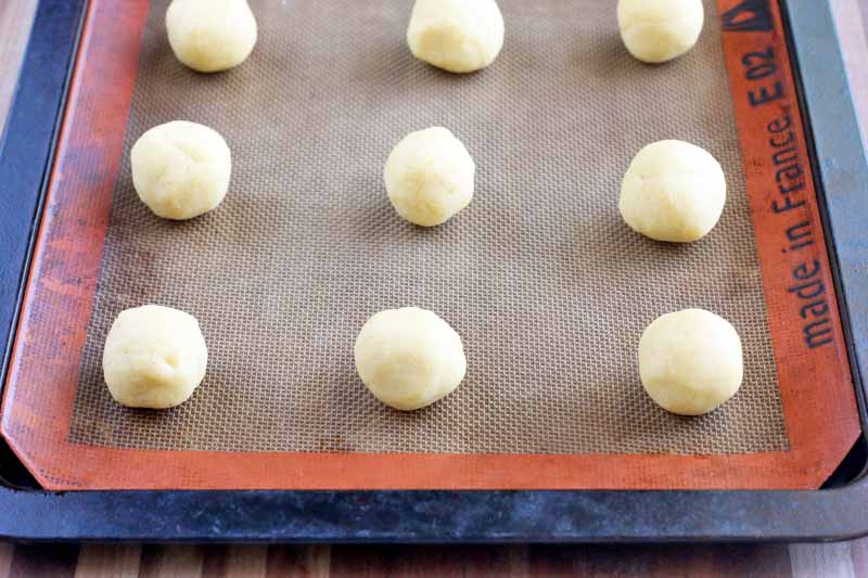 Balls of cornmeal butter cookie dough are arranged in rows on a brown and orange Silpat liner on top of a metal sheet pan.