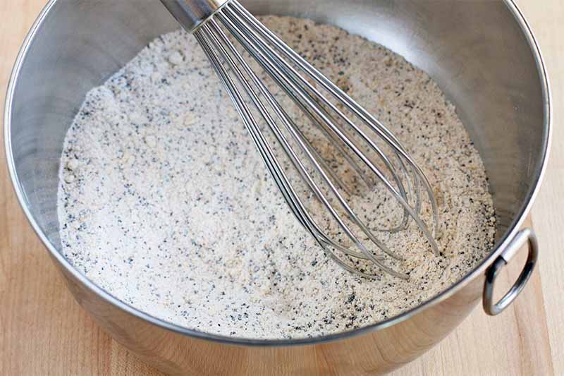 A whisk mixes a dry flour mixture that is speckled with poppy seeds in a stainless steel bowl, on a beige countertop.