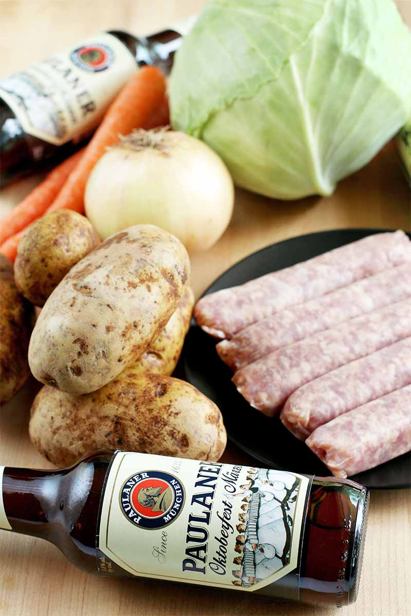 Potatoes, carrots, an onion, a green cabbage, two bottles of beer, and a plate topped with five German sausages, on a beige background.