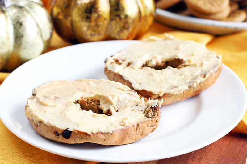 A cinnamon raisin bagel spread with pale orange pumpkin cream cheese is on a white plate, on a surface topped with a yellow cloth, with silver and gold decorative pumpkins and a plate of cookies in the background.