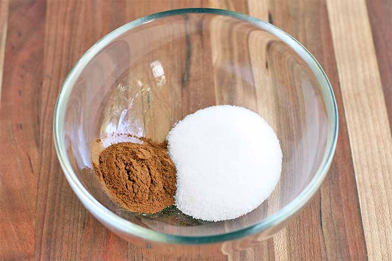 A pile of cinnamon is to the left of a larger pile of granulated sugar, in a clear glass bowl on a brown wood surface.