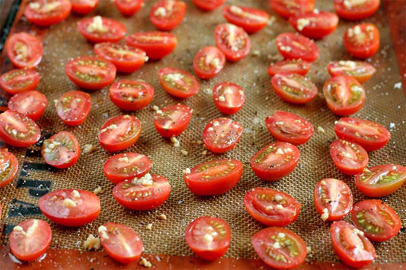 Halved grape tomatoes sprinkled with garlic, salt, and pepper, arranged in a single layer on a silicone Silpat pan liner.