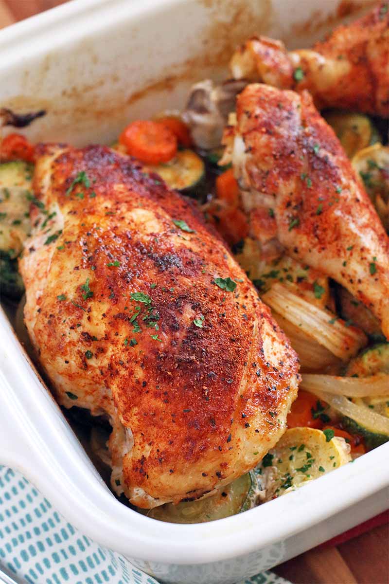 Closeup of a white ceramic baking dish of oven-roasted chicken and vegetables, coated with a red-orange spice mixture.