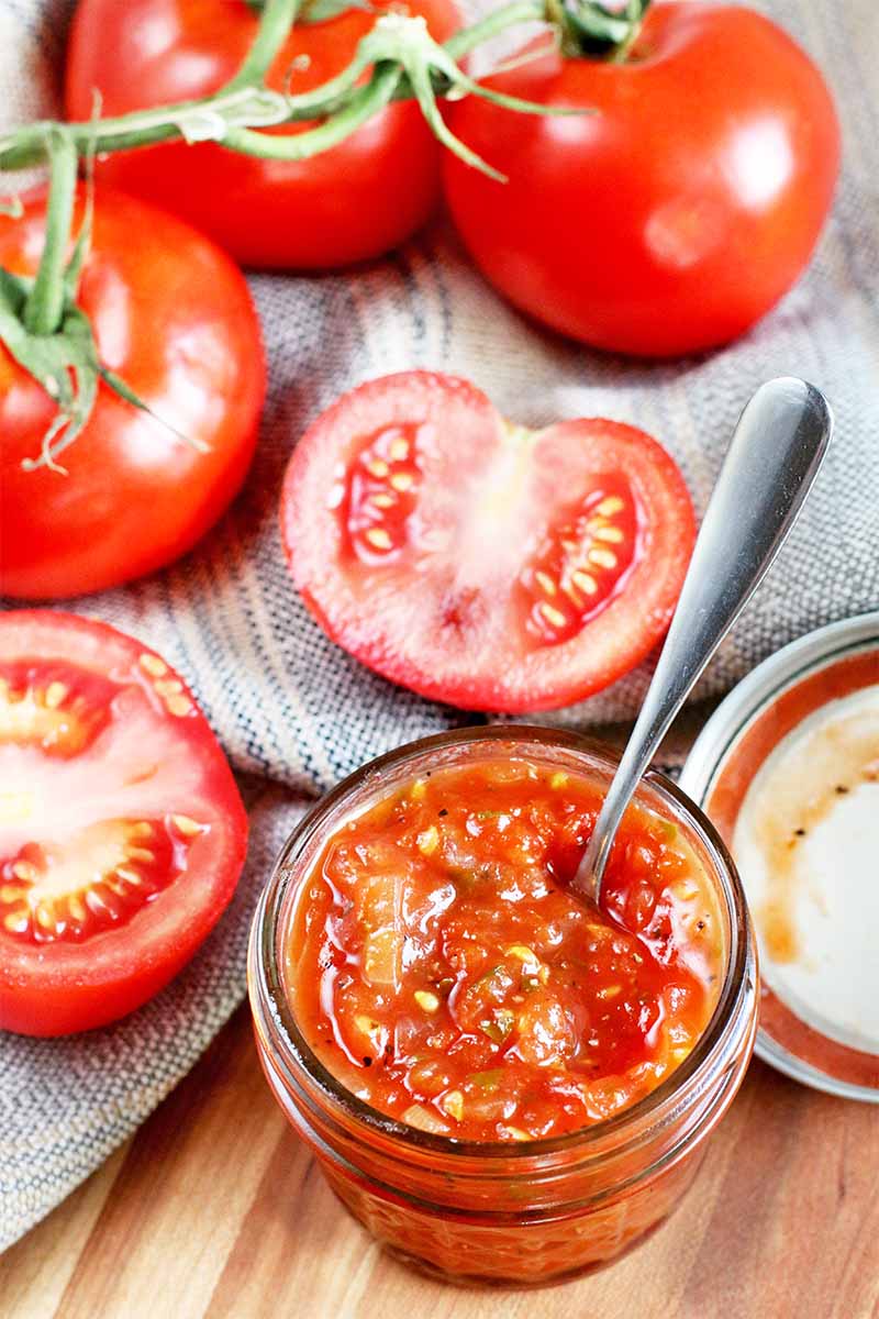 Halved and whole fresh tomatoes and homemade jam in a small glass jar, with a metal lid and a silver spoon, on a gray cloth on top of a wood surface.