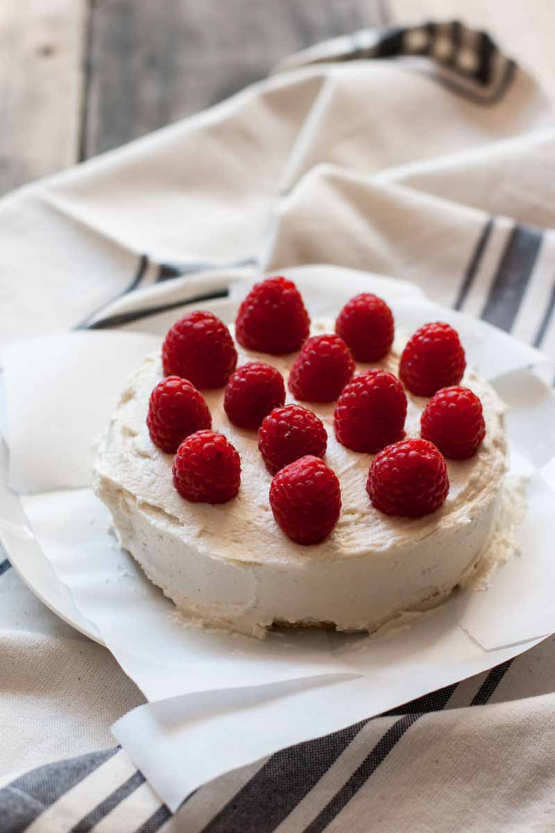 Vertical image of one round layer covered in frosting and topped with raspberries on a blue-striped towel.