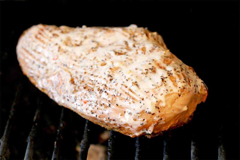 A spice-rubbed chicken breast is spread with white barbecue sauce, cooking on a black metal grill grate.