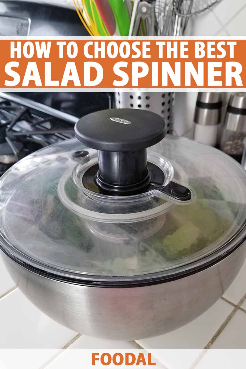 OXO stainless steel, acrylic, and black plastic salad spinner on a white tile countertop next to a gas stove, with a stainless canister of utensils and salt and pepper grinders in the background, with orange and white text.