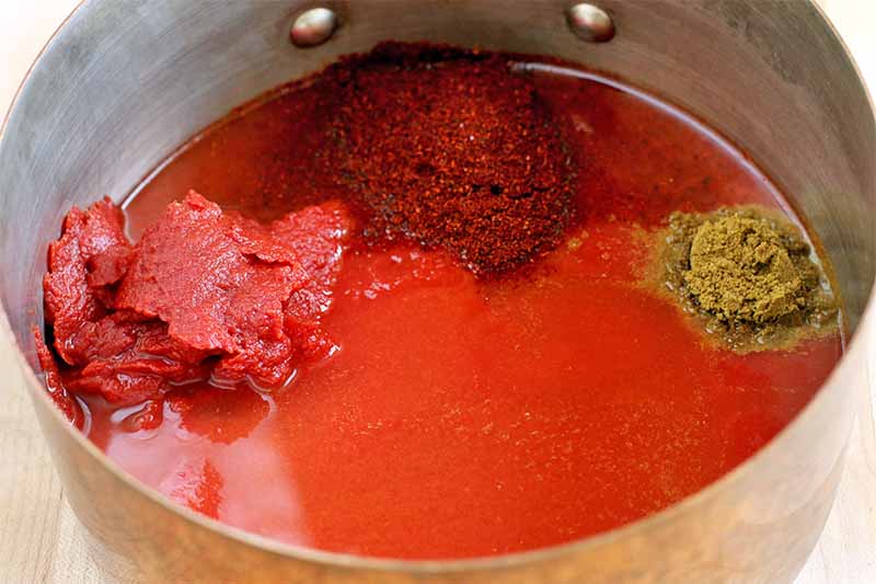 Red hot sauce and piles of tomato paste, chili powder, and ground cumin are in the bottom of a saucepan, on a beige background.