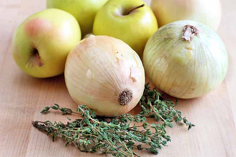 Three onions, three green apples with a pink blush, and several sprigs of thyme are arranged on a beige wood surface.
