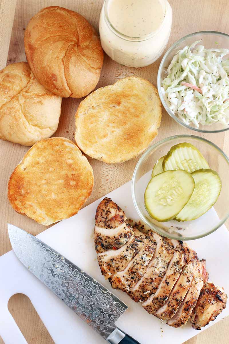 Top-down shot of a sliced spice-rubbed grilled chicken breast on a white plastic cutting board with a chef's knife, toasted kaiser rolls, a glass jar of white sauce, and two small glass bowls of coleslaw and pickles, on a beige background.