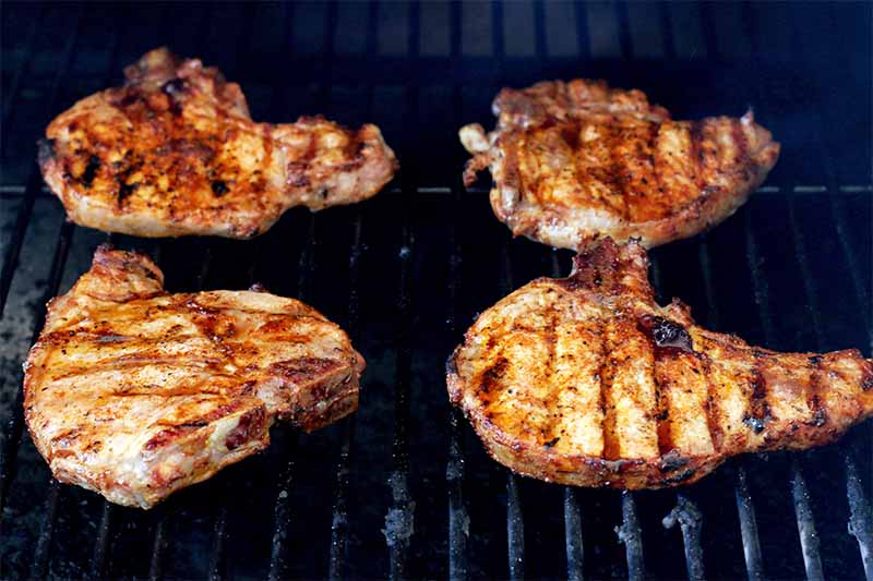 Four spice-rubbed pork chops cooking on the grate of a black barbecue grill.