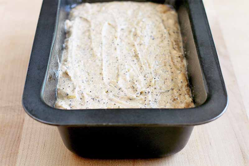 A black metal baking pan is filled with quick bread batter, on a beige countertop.