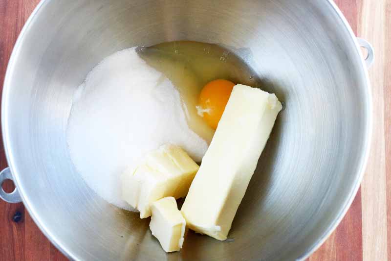 Whole and partial sticks of butter are in the bottom of a stainless steel mixing bowl with a raw egg without the shell, and white sugar.