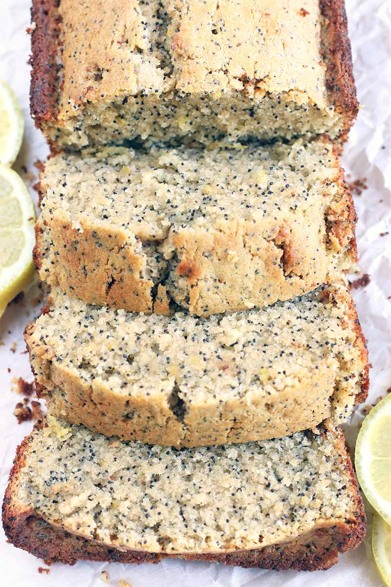 Closely cropped vertical shot of three slices and half a loaf of lemon poppy seed bread, on a piece of white parchment paper with decorative lemon slices.