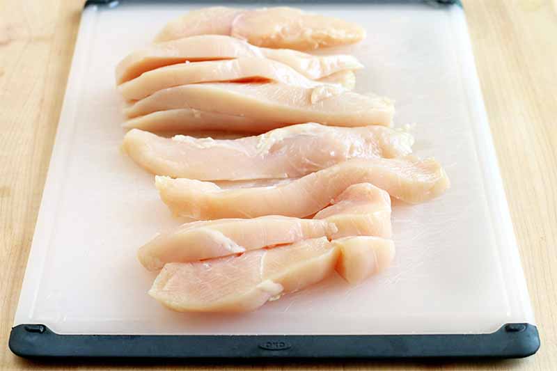 Sliced chicken breast arranged in a column on a black and white plastic cutting board, on a beige countertop.