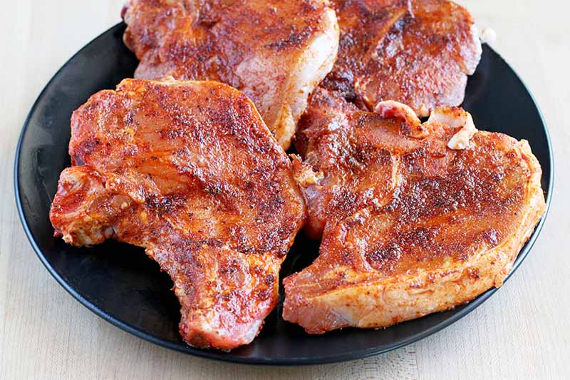 Four bone-in pork chops on a black plate, rubbed with a red spice mixture, on a beige background.