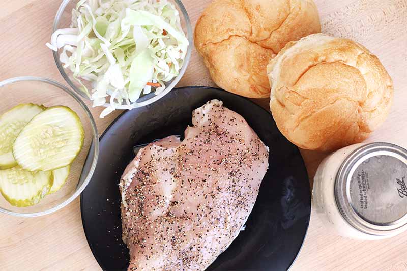 Top-down shot of a raw chicken breast rubbed with spices on a small black plate, surrounded by small glass dishes of pickle slices and shredded cabbage, two kaiser rolls, and a mason jar filled with a white liquid, on a beige surface.