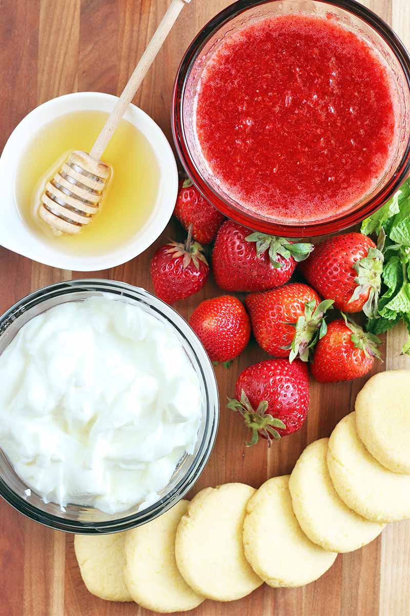 Top-down shot of a small white bowl of honey with a wooden dipper, glass bowls of strawberry coulis and Greek yogurt, a fanned out pile of homemade lemon cornmeal cookies, and fresh strawberries, on a brown wood surface.