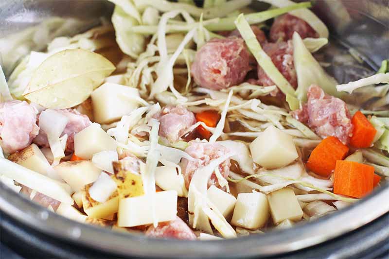 Closeup shot of chunks of potato and carrot, shredded cabbage, sliced bratwurst, and a bay leaf, in a slow cooker.