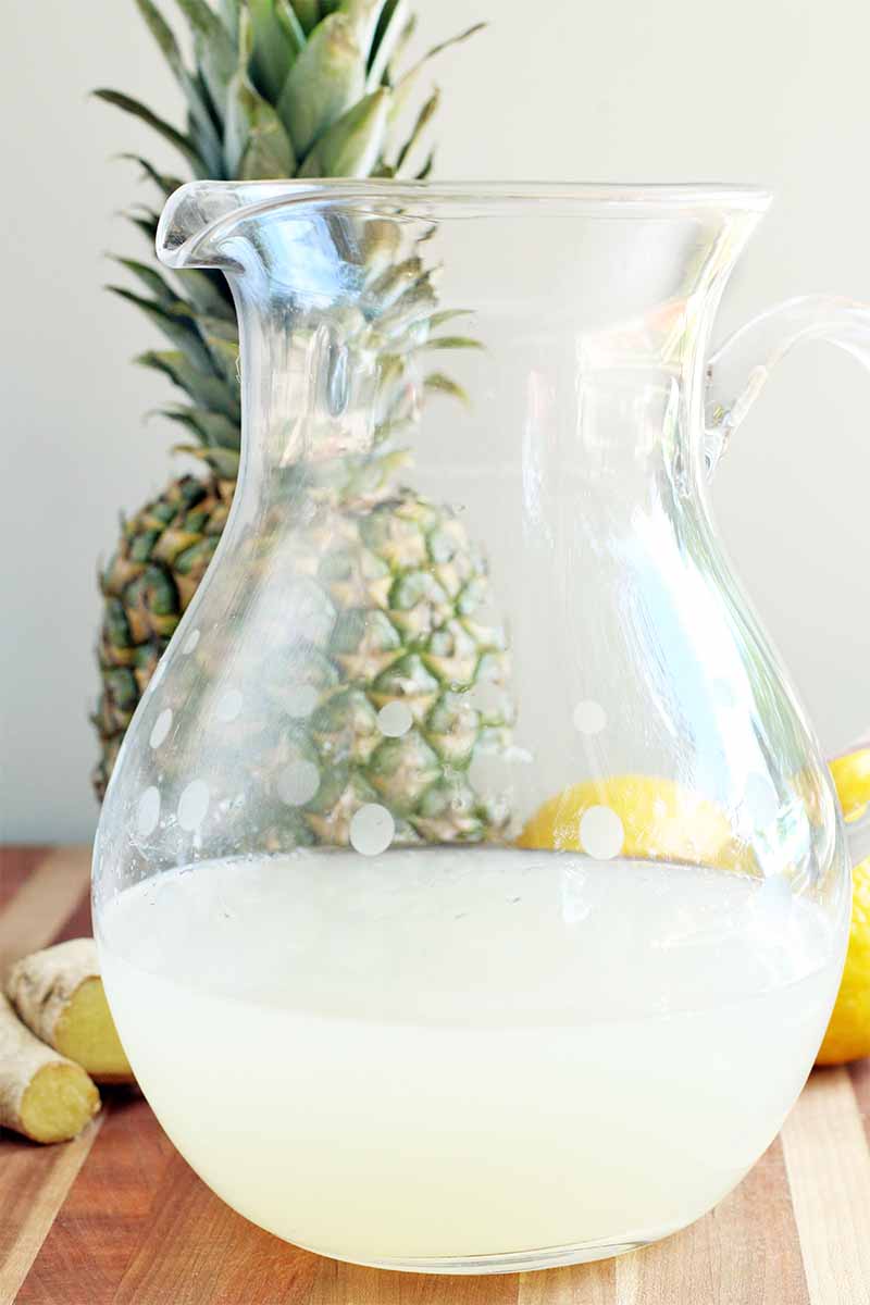 A glass pitcher filled about 1/3 of the way with a pale yellow lemonade, with a whole pineapple, ginger, and lemons in the background, on a wood surface against a gray backdrop.