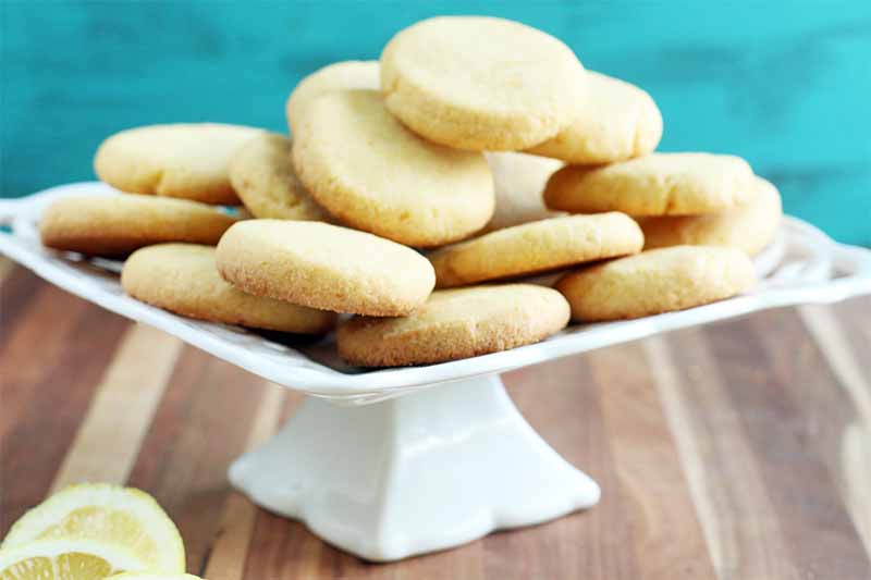 Lemon cookies are arranged on a white ceramic pedestal serving platter, with sliced yellow citrus on a wood surface, with an aqua background.
