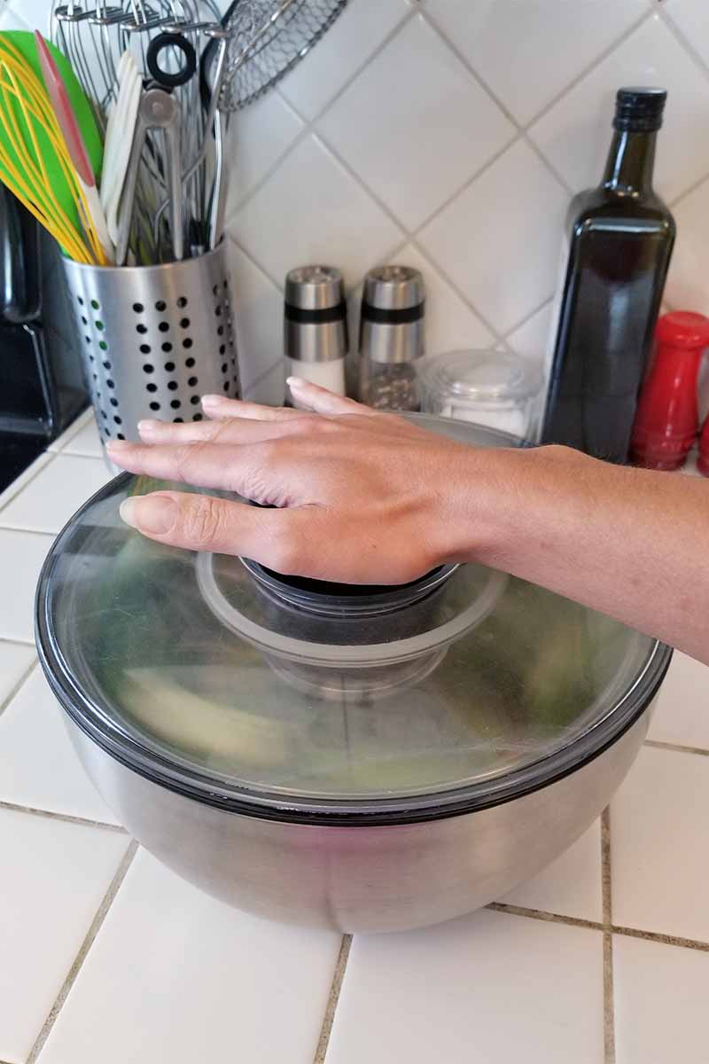 A hand presses down on the top plunger that makes a salad spinner bowl rotate to dry the greens, with an acrylic top and stainless steel base, on a white tile kitchen counter.