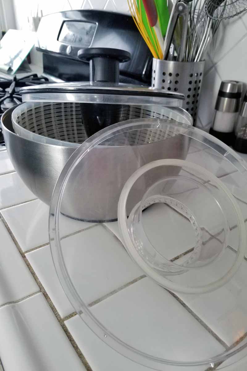 A clear acrylic inner ring rests against the stainless steel outer bowl of a salad spinner, with a plastic net-like bowl inside, and a lid on top with spinning central hub and components made of black plastic, on a white tile countertop.