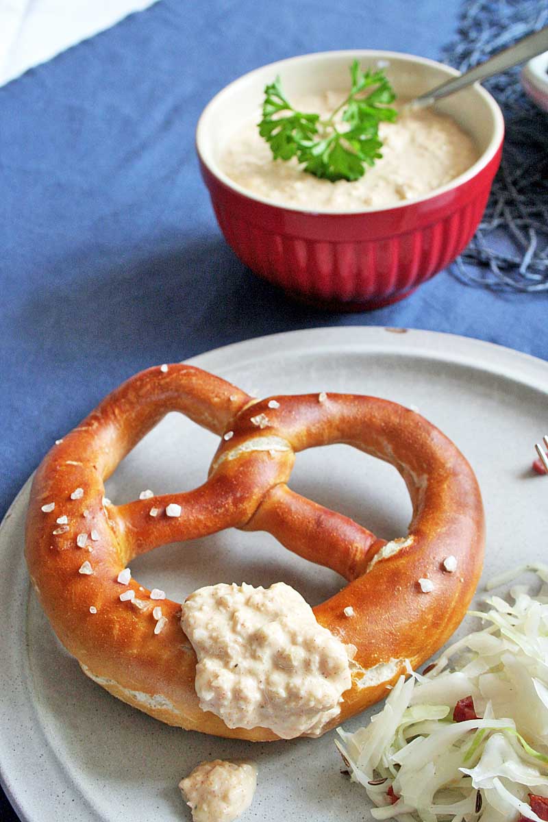 Close up of a German lye pretzel with a dab of Obatzda cheese dip on it. A bowl of the dip is in the background. Sitting on a medium-blue tablecloth.