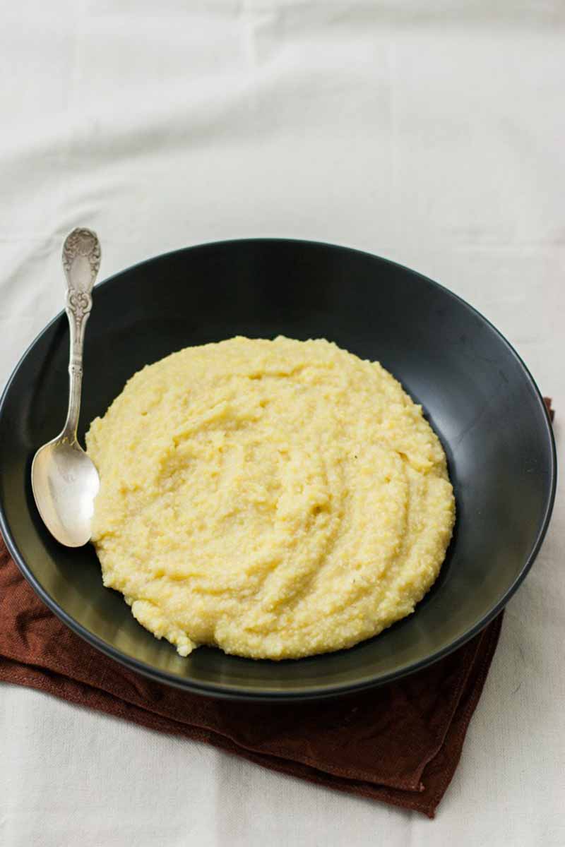 Vertical image of a black bowl of polenta with a metal spoon on a napkin.