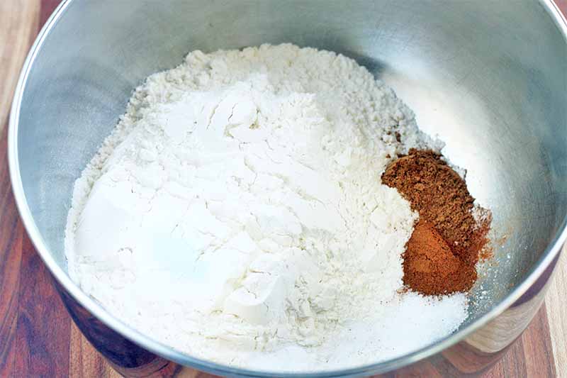Flour and cinnamon are in the bottom of a stainless steel mixing bowl, on a wood table.