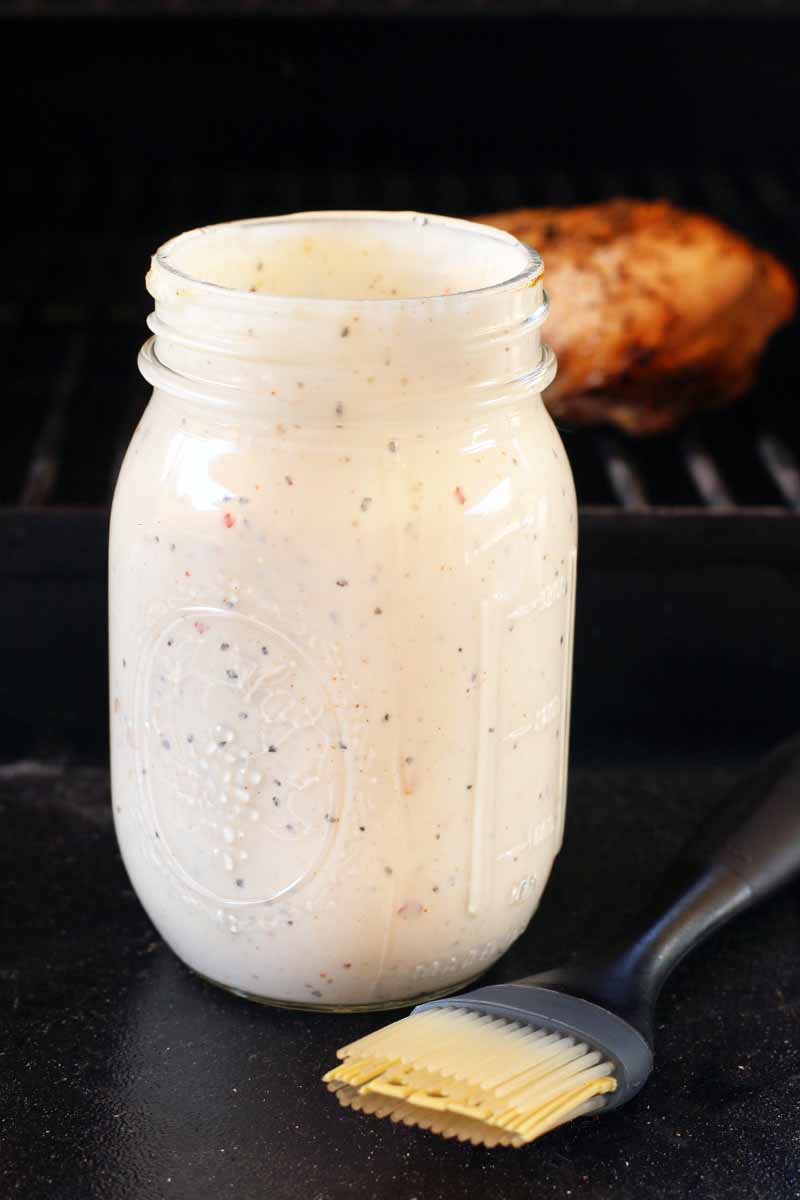 A Mason jar of white barbecue sauce and a black and straw-colored silicone brush are on the foreground, with a piece of spice-rubbed chicken on a metal grate in the background, on a black surface against a black backdrop.