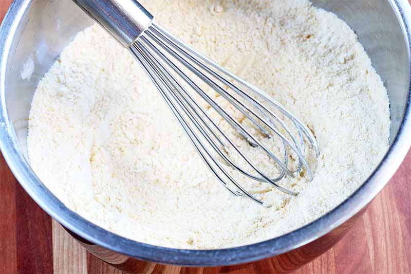 A silver wire whisk is stirring a dry flour mixture in a stainless steel mixing bowl, on a wood countertop.