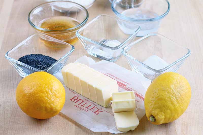 Sliced butter on a waxed paper wrapper with two lemons and small square and round glass dishes of poppy seeds, agave syrup, salt, baking soda, and other ingredients, on a beige countertop.