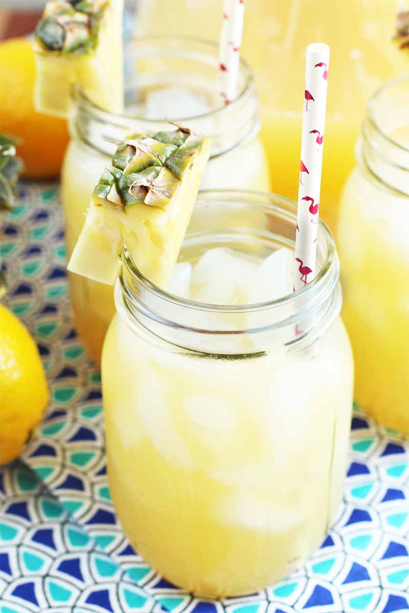 Vertical image of three mason jars of tropical lemonade, with paper straws and pineapple garnish, with a whole lemon to the left of the frame, on a dark and light blue patterned cloth surface.