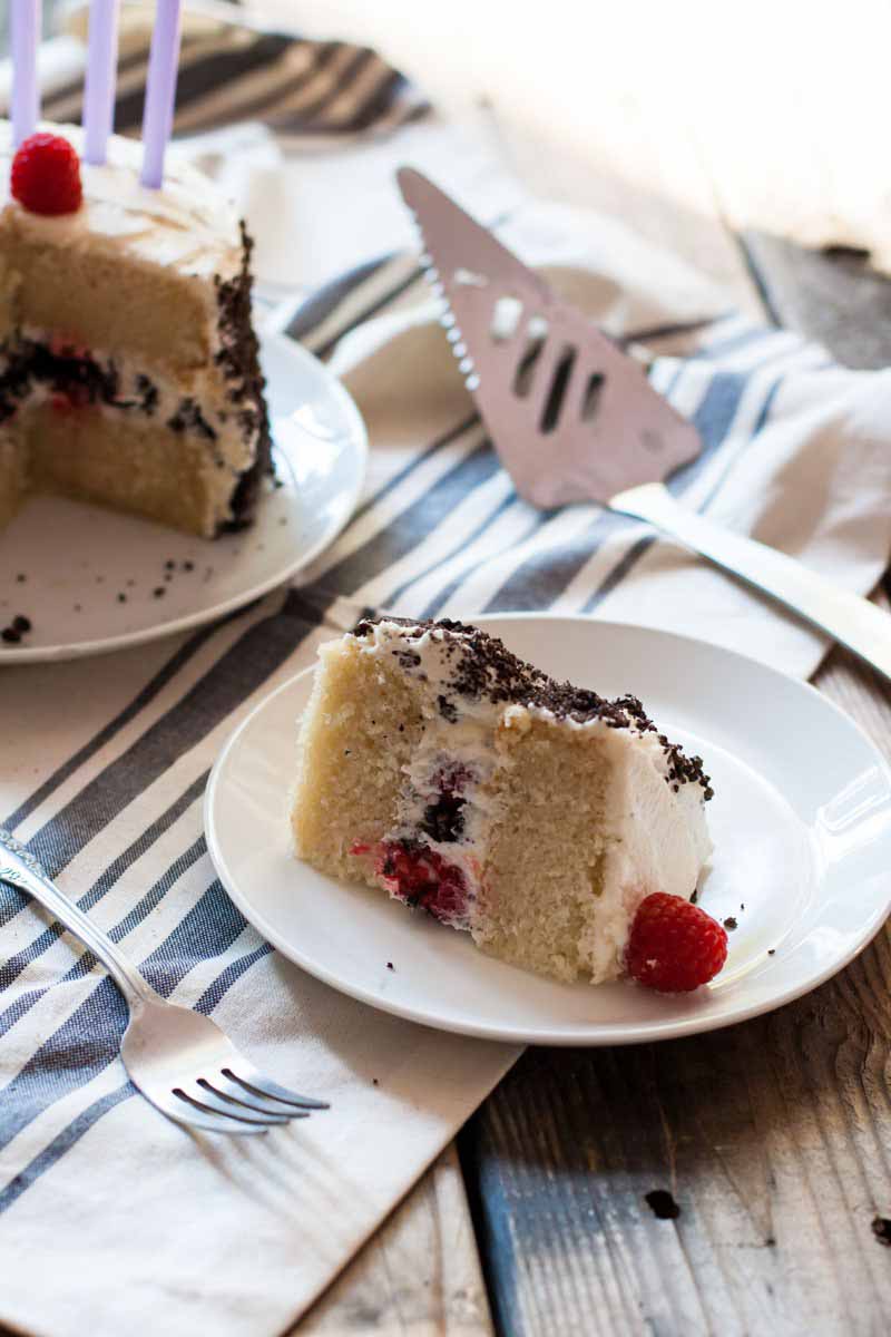 Vertical image of a small slice of cake with a raspberry on a white plate, with a fork and serving knife.