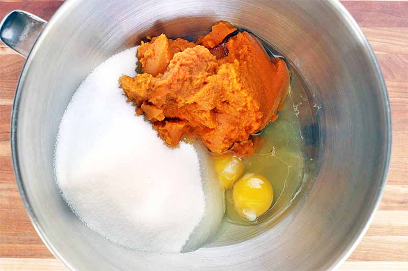 Orange pumpkin puree, two eggs without the shell, and a pile of sugar rest at the bottom of a stainless steel stand mixer bowl, on a light brown surface.