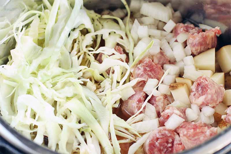 Shredded cabbage with pieces of raw potato and bratwurst, in the bowl of a slow cooker.