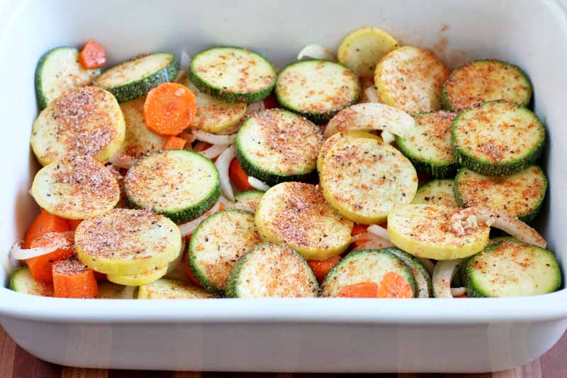 Sliced yellow summer squash, zucchini, onions, and carrots, in a rectangular ceramic baking dish, sprinkled with a red spice mixture.