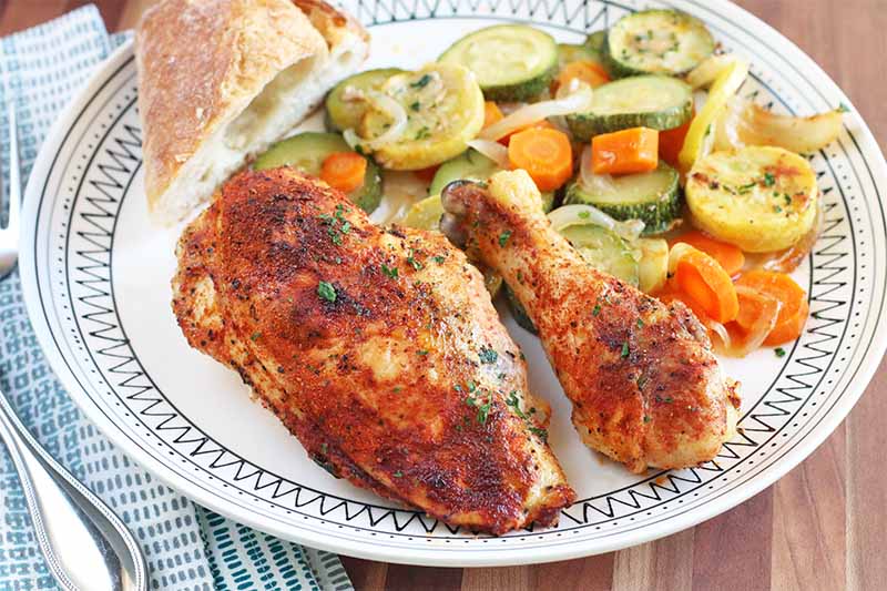 Roasted spiced chicken breast and a drumstick on a dinner plate with squash and root vegetables, and a piece of bread, beside silverware on a blue and white cloth napkin, on a brown wood table.