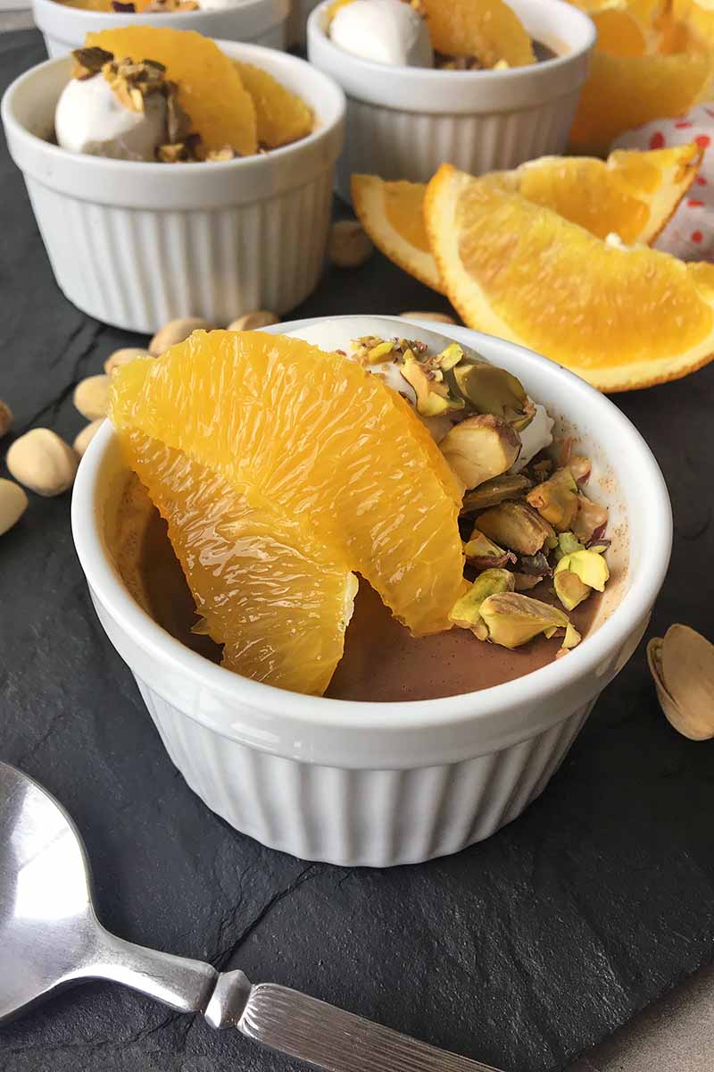 Vertical image of white ramekins with a dessert garnished with segmented oranges and pistachios, with a metal spoon on the side.