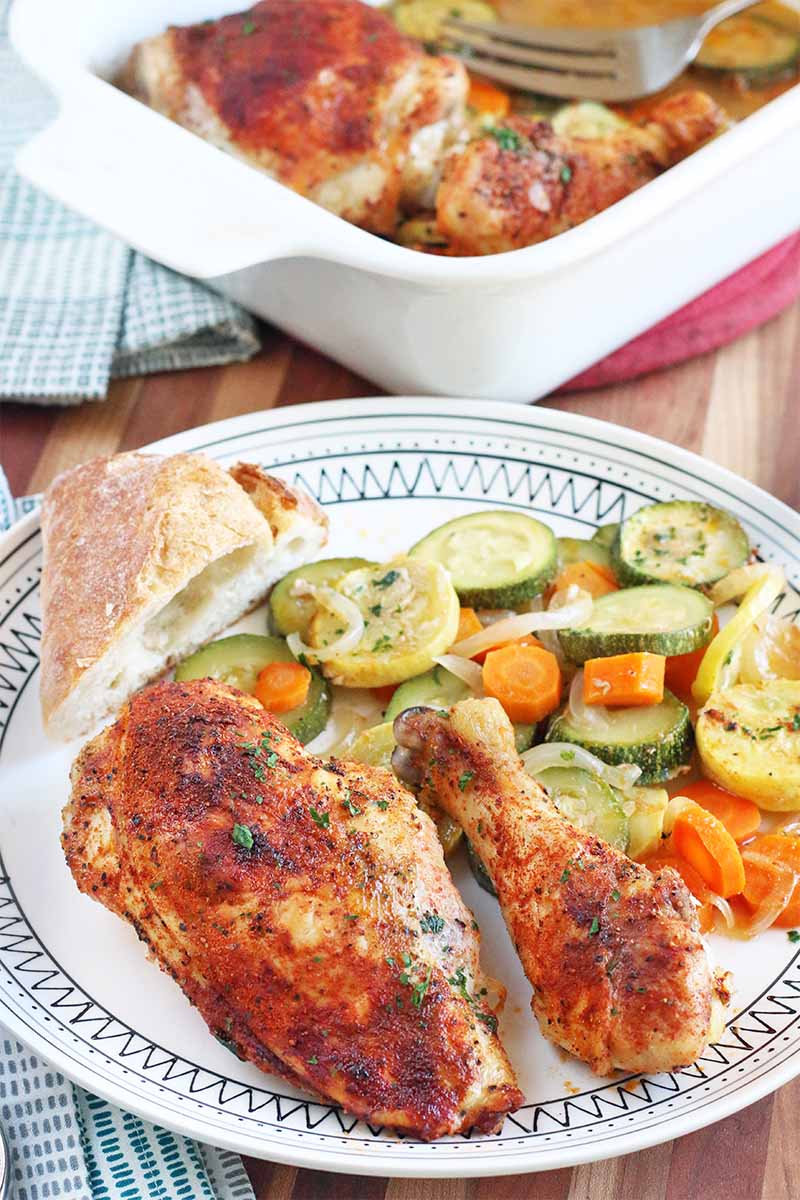 Vertical image of a white baking dish and a black and white patterned dinner plate of roasted chicken coated with a red spice mixture and sliced carrots, onions, and summer squash, with a triangle of bread on the edge of the plate, and a folded cloth napkin on a brown wood surface.