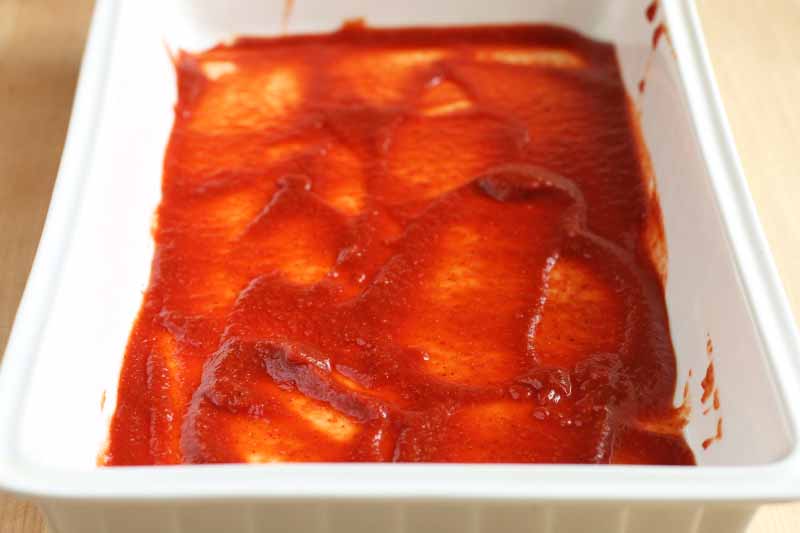Red sauce is spread in the bottom of a white, rectangular baking dish, on a beige surface.