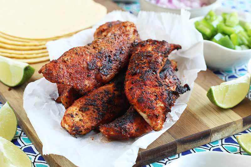 Blackened Cajun chicken tenders on a crumpled piece of white parchment paper, on a wood cutting board with lime wedges and a stack of corn tortillas, with small ceramic bowls of chopped green pepper and onion, on a dark and light blue patterned cloth surface.