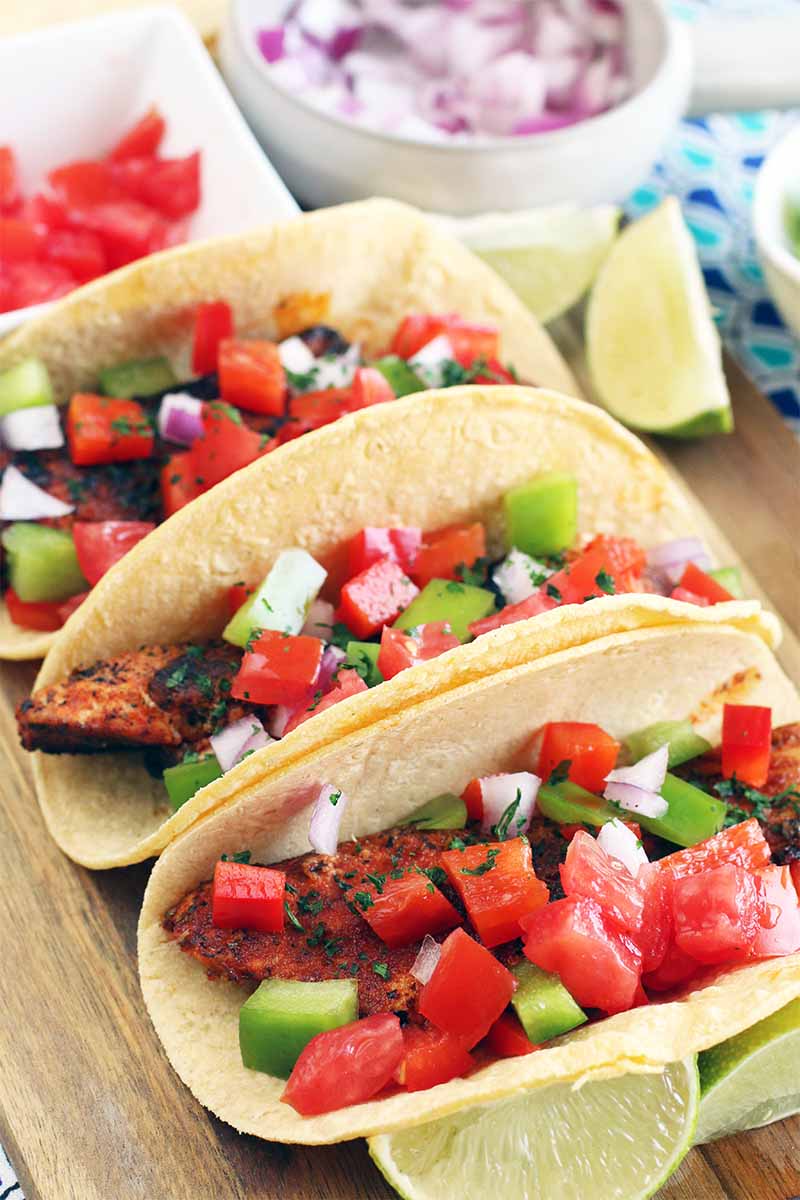 Three Cajun chicken tacos garnished with chopped vegetables and surrounded by lime wedges on a wood cutting board, with small white bowls of chopped tomato and purple onion in the background.