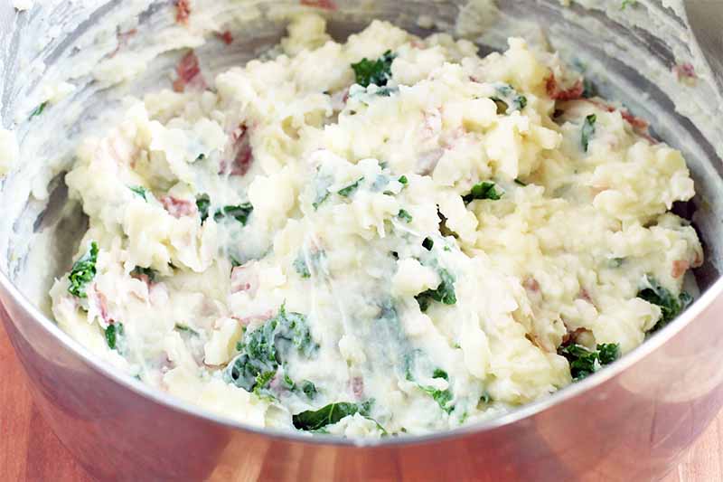 A stainless steel bowl of mashed red-skinned potatoes with green kale, on a brown wood surface.