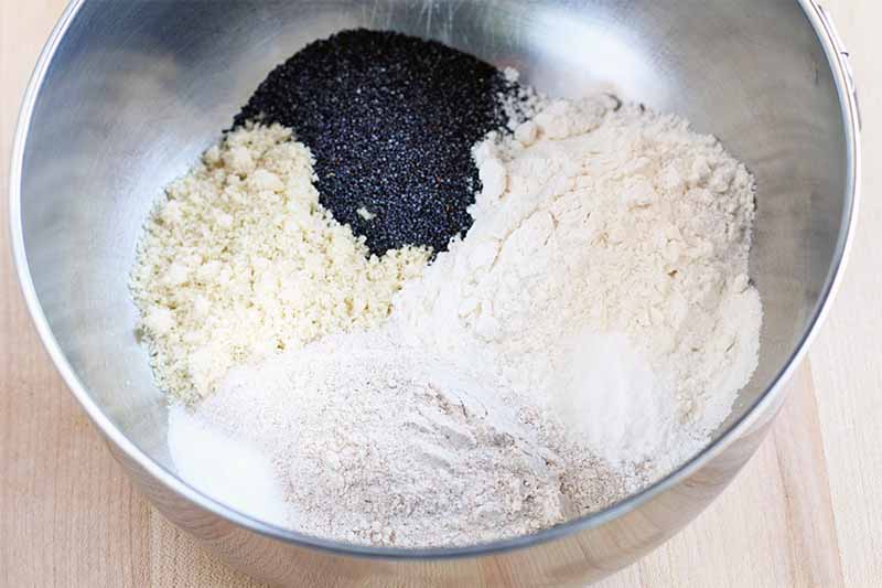 Piles of flour and poppy seeds in the bottom of a stainless steel mixing bowl, on a beige countertop.
