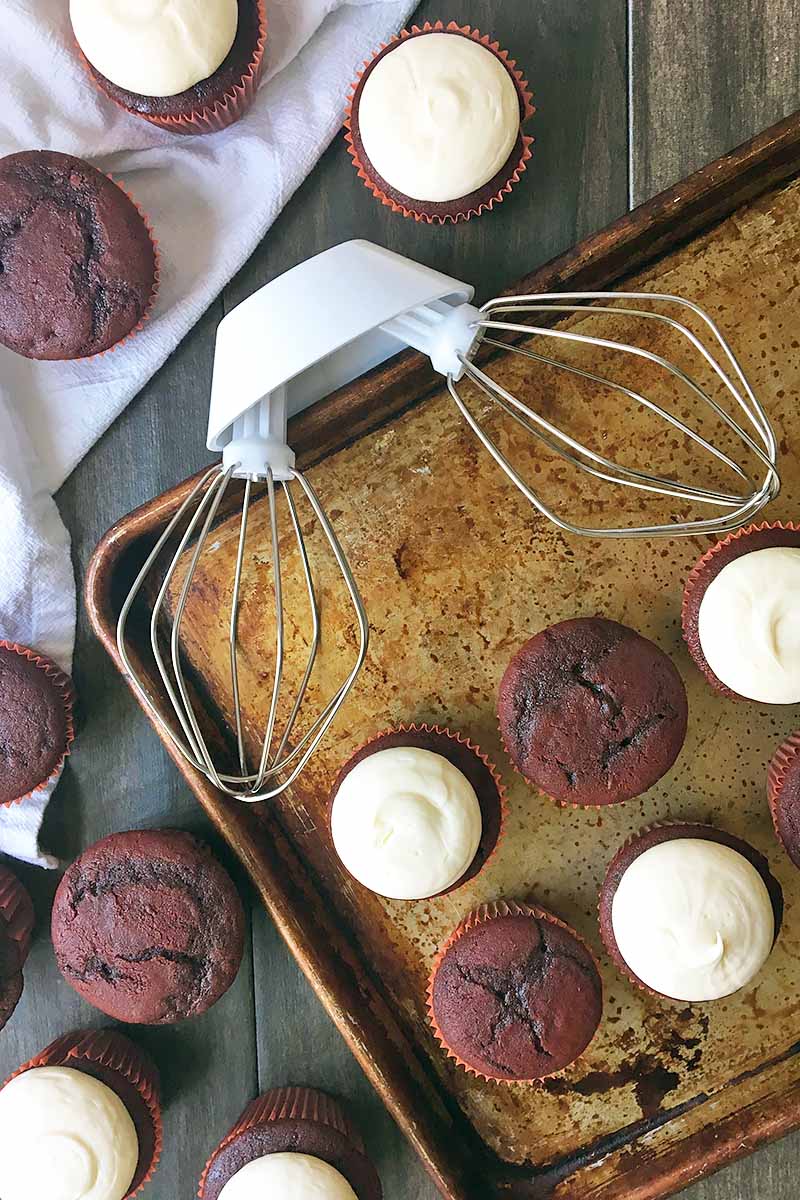 Vertical image of dual whisks next to a sheet pan with cupcakes.