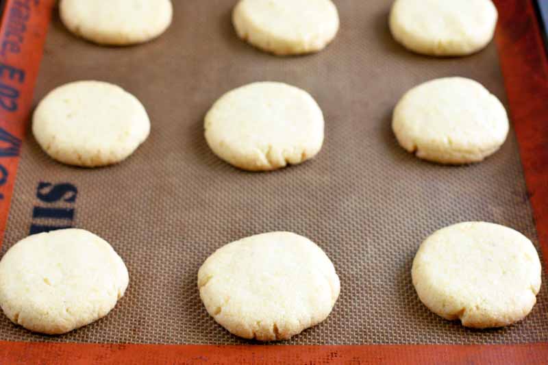 Round, pale off-white cookies with cracked edges on a silicone Silpat liner, arranged in rows.