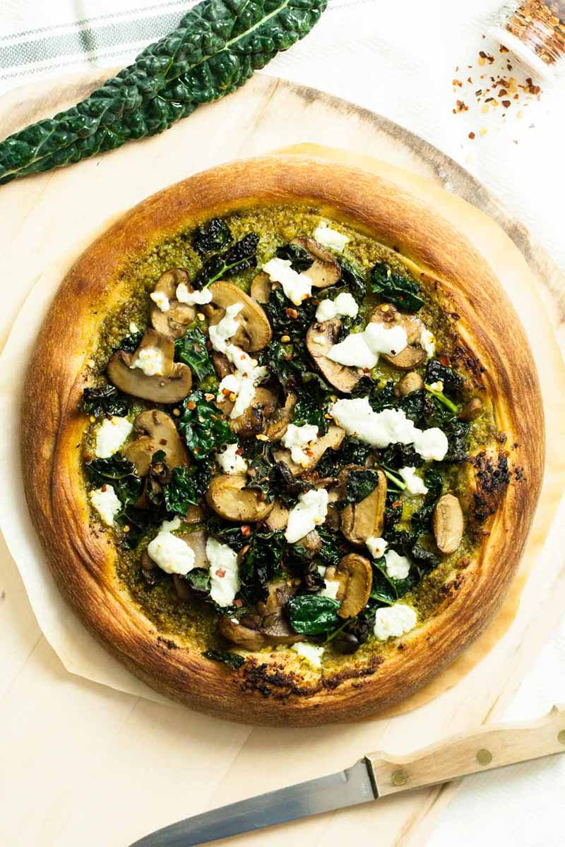 Vertical top-down image of a whole pizza with greens, goat cheese, and mushrooms.
